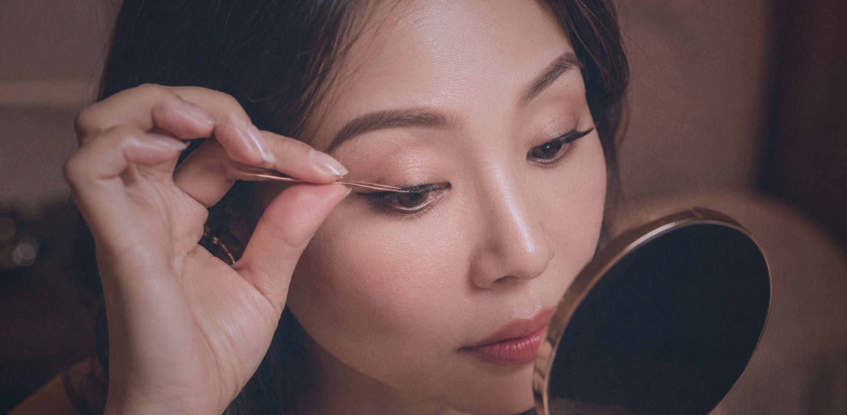 Singaporean woman applying press-on lashes with a precision applicator while looking into a mirror, showing a simple no-glue DIY lash extension routine.