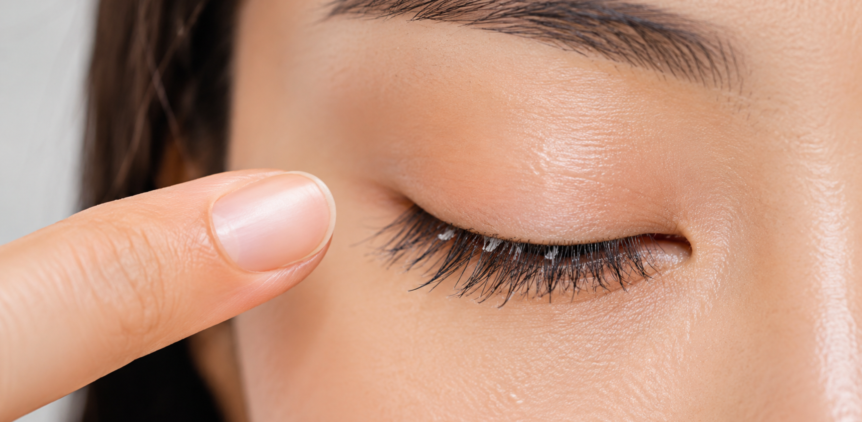 Close-up of an Asian woman’s closed eye with a finger pointing to white residue on natural lashes caused by false lash glue buildup.
